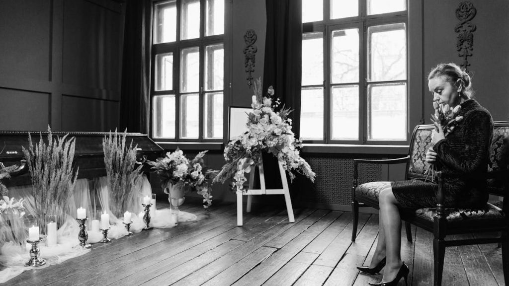 Grayscale funeral scene showing a woman in mourning by a coffin and floral arrangements.